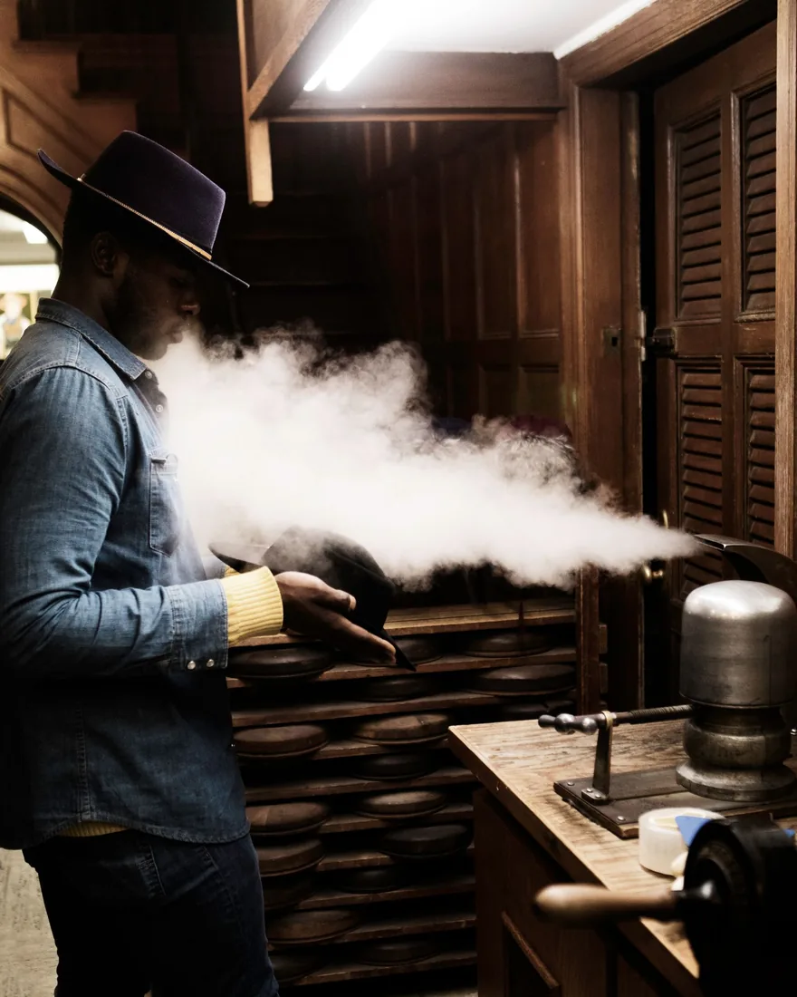 Formado de un sombrero con vapor en un taller de sombrerería, junto a una estantería con hormas de madera apiladas