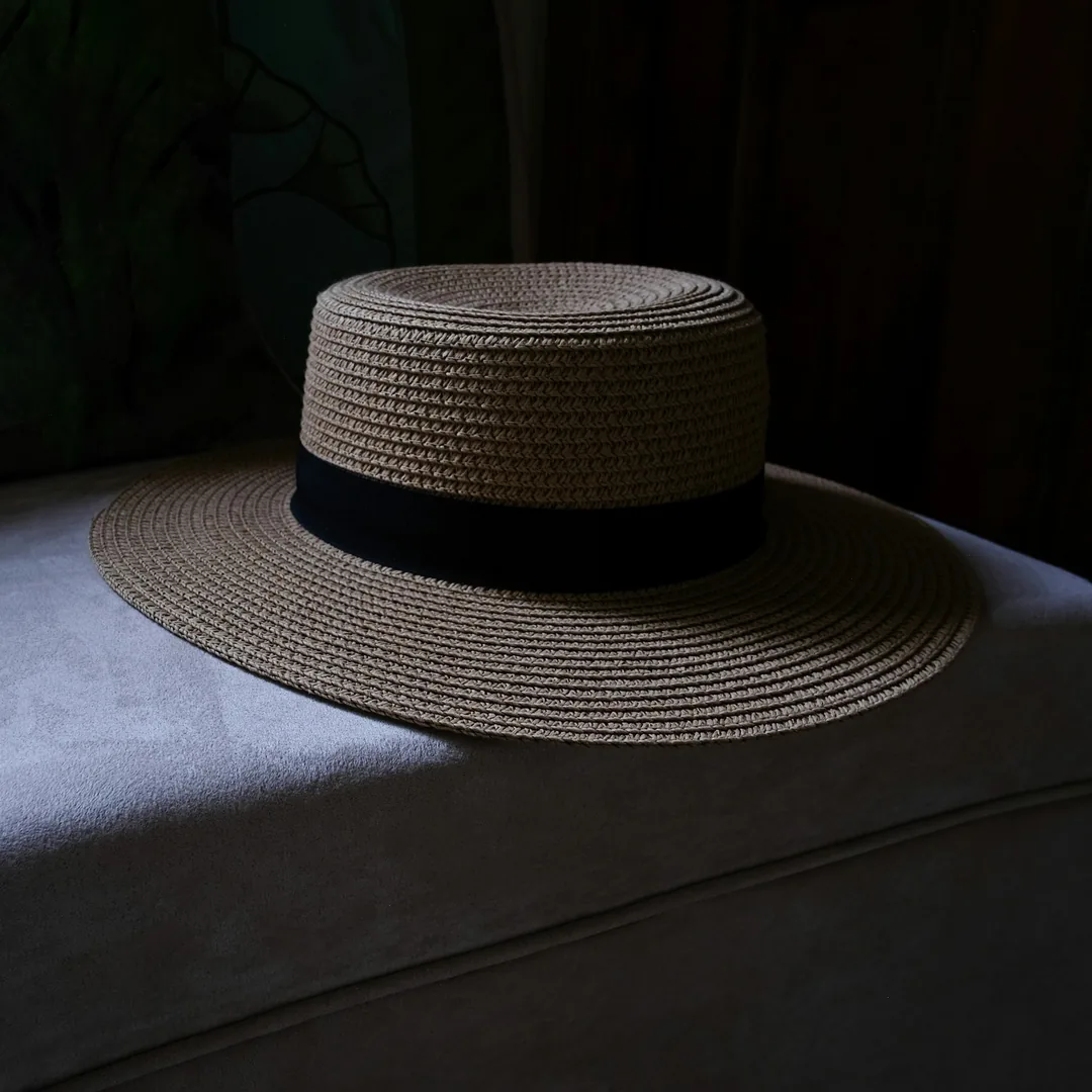 Sombrero de paja trenzada de copa plana y ala ancha, con cinta oscura en la base de la copa, apoyado sobre una tela clara